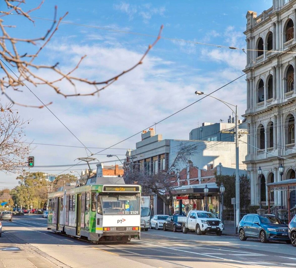 A tram is seen traversing a street lined with parked cars and a mix of commercial buildings. The architecture features classic and modern designs, with trees planted along the footpath. A clear blue sky with scattered clouds is visible overhead.