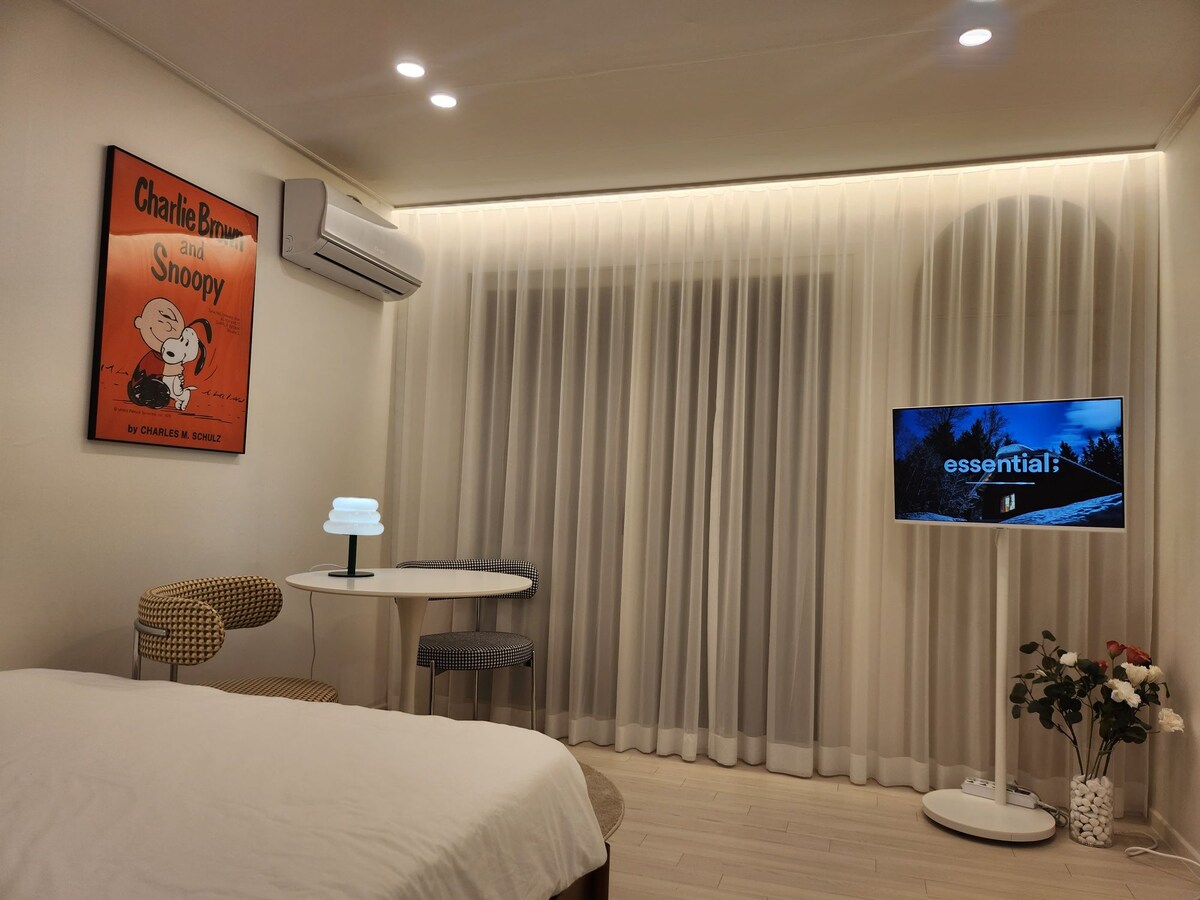 A cozy bedroom features a spacious bed with soft linens and a wall mounted television. A bright lamp sits on a small round table beside a patterned chair. The room is illuminated by soft lighting and adorned with a curtain that allows natural light to filter in.