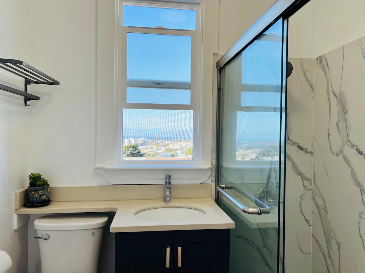 A bright bathroom features a large window offering a view of the surroundings. A modern glass shower door stands adjacent to a marble-topped vanity, which is paired with a dark cabinet. A small plant adds a touch of greenery, and essentials are organized on a wall-mounted shelf.