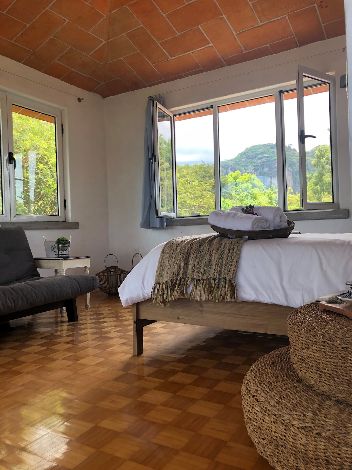 A light-filled room features large open windows offering views of the surrounding trees and hills. A spacious bed with neutral-toned bedding is complemented by a textured throw. A cozy seating area includes a gray chair, while a woven ottoman adds natural texture to the space.