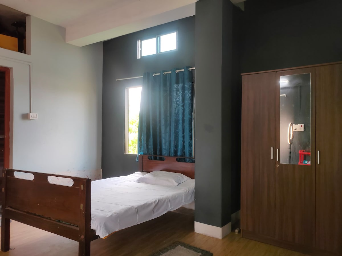 A spacious sleeping area is highlighted by a wooden bed frame and a neatly made bed. Natural light enters through two small windows, with dark curtains partially drawn. A wardrobe with mirrored doors stands beside the bed, adding functionality to the room.