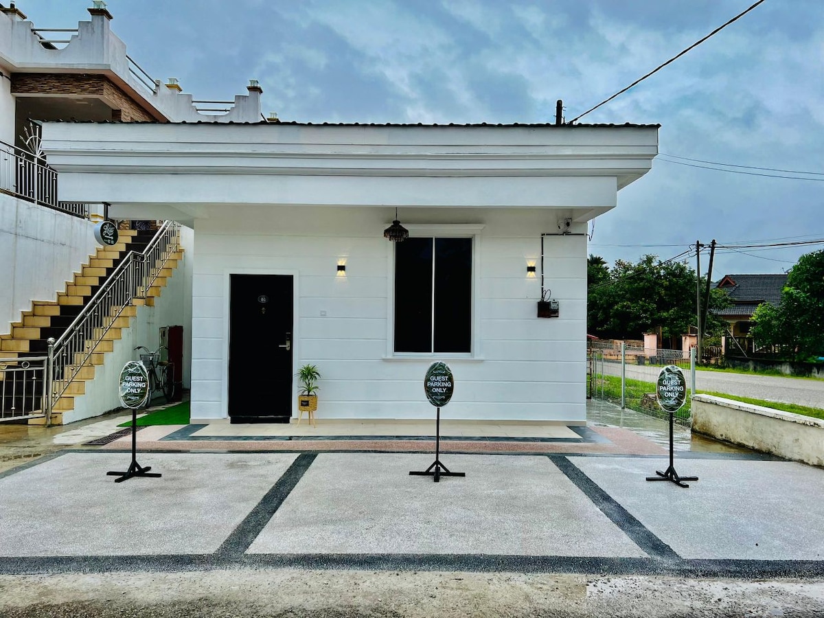 The exterior of the guest house is presented with a clean white facade and a single door. A large window is centered on the front, allowing natural light to enter. Signage is placed on stands along the paved area, providing information for guests.