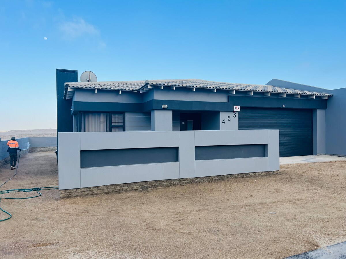A modern single-story house is presented, featuring a sleek façade with grey coloring. A spacious driveway leads to a covered garage. An entrance is visible, framed by a low wall. A person is seen watering the garden in the background.