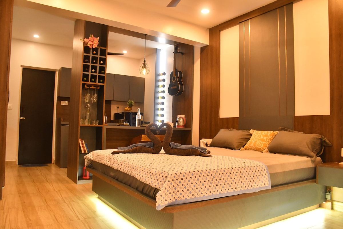 A spacious sleeping area features a large bed adorned with decorative pillows and a heart-shaped towel arrangement. The room's wooden accents complement the modern kitchenette, while soft lighting creates a cozy ambiance. A guitar is displayed on the wall, enhancing the inviting atmosphere.
