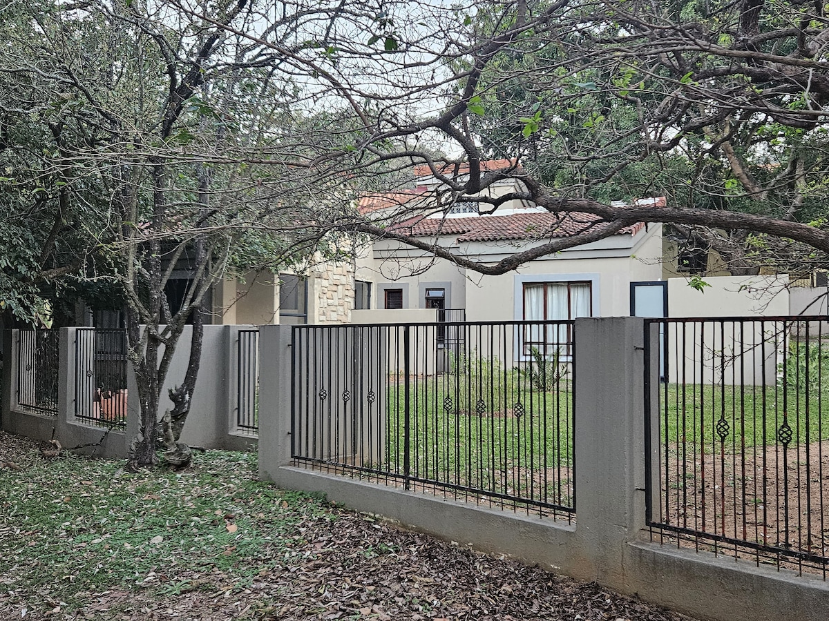 A tranquil outdoor scene features a modest home surrounded by greenery and mature trees. A black wrought iron fence provides a clear boundary, while leaves cover the ground. Large windows are visible, allowing for natural light to enter the interior.