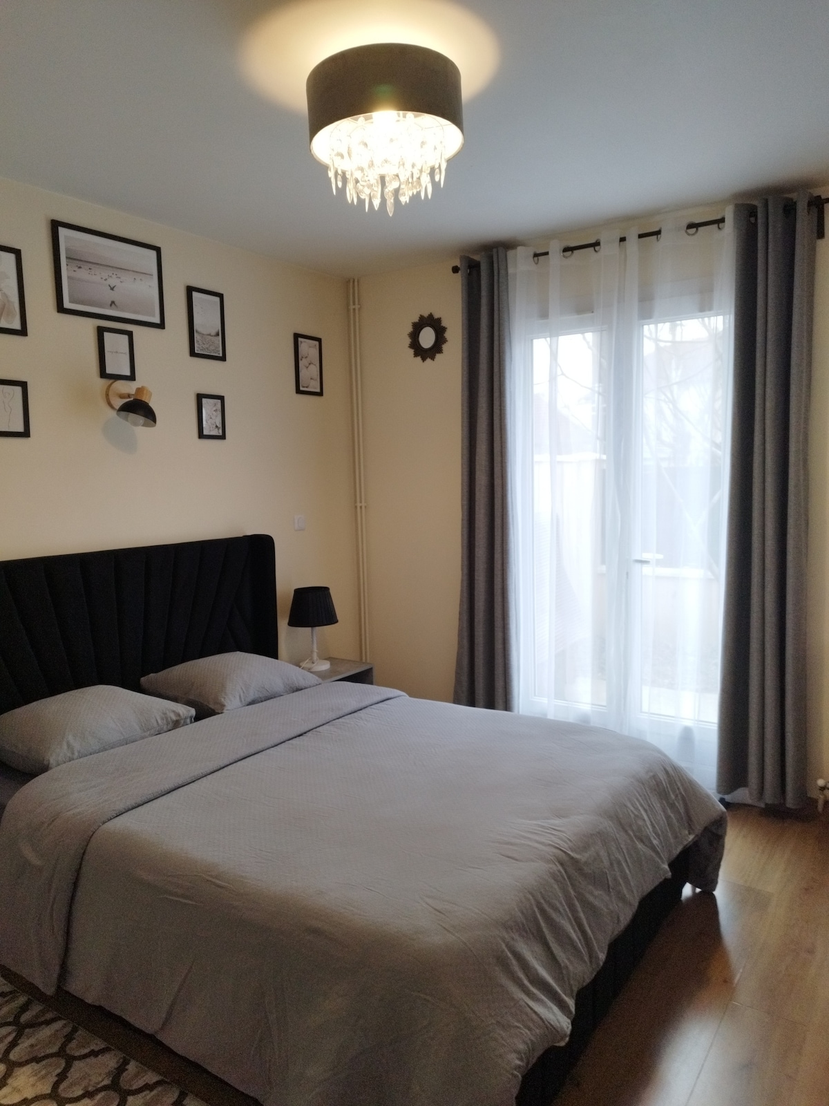 A neatly arranged bedroom features a spacious bed with a soft grey coverlet and two plush pillows. A pair of framed pictures decorates the wall, and a small table lamp complements the decor. Natural light filters through a pair of sheer curtains covering the window.
