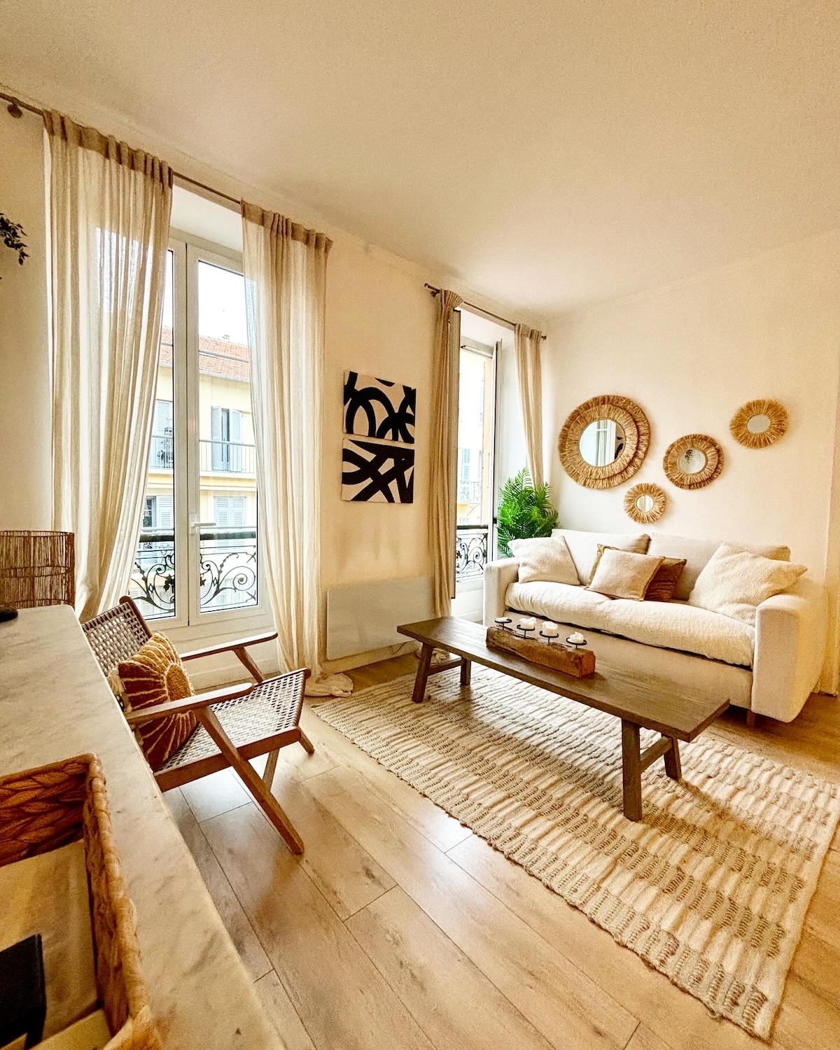 The living area features large windows dressed with light, flowing curtains, allowing natural light to fill the space. A comfortable white sofa is positioned next to a wooden coffee table, which is centered on a woven area rug. Decorative wall accents complement the ambiance.
