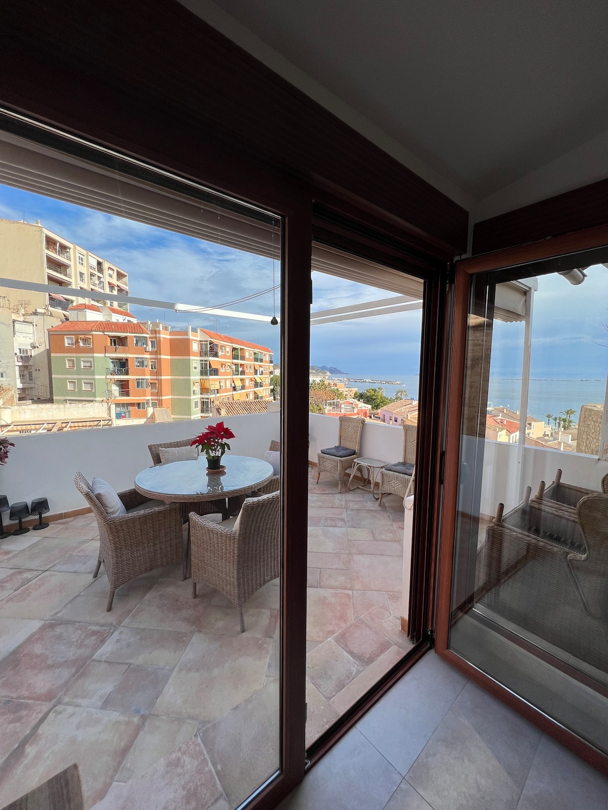 A terrace is visible through sliding glass doors, featuring a round table surrounded by four wicker chairs. The tiled floor complements the scenic view of colorful buildings and the sea in the background. Natural light brightens the space, enhancing the inviting atmosphere.