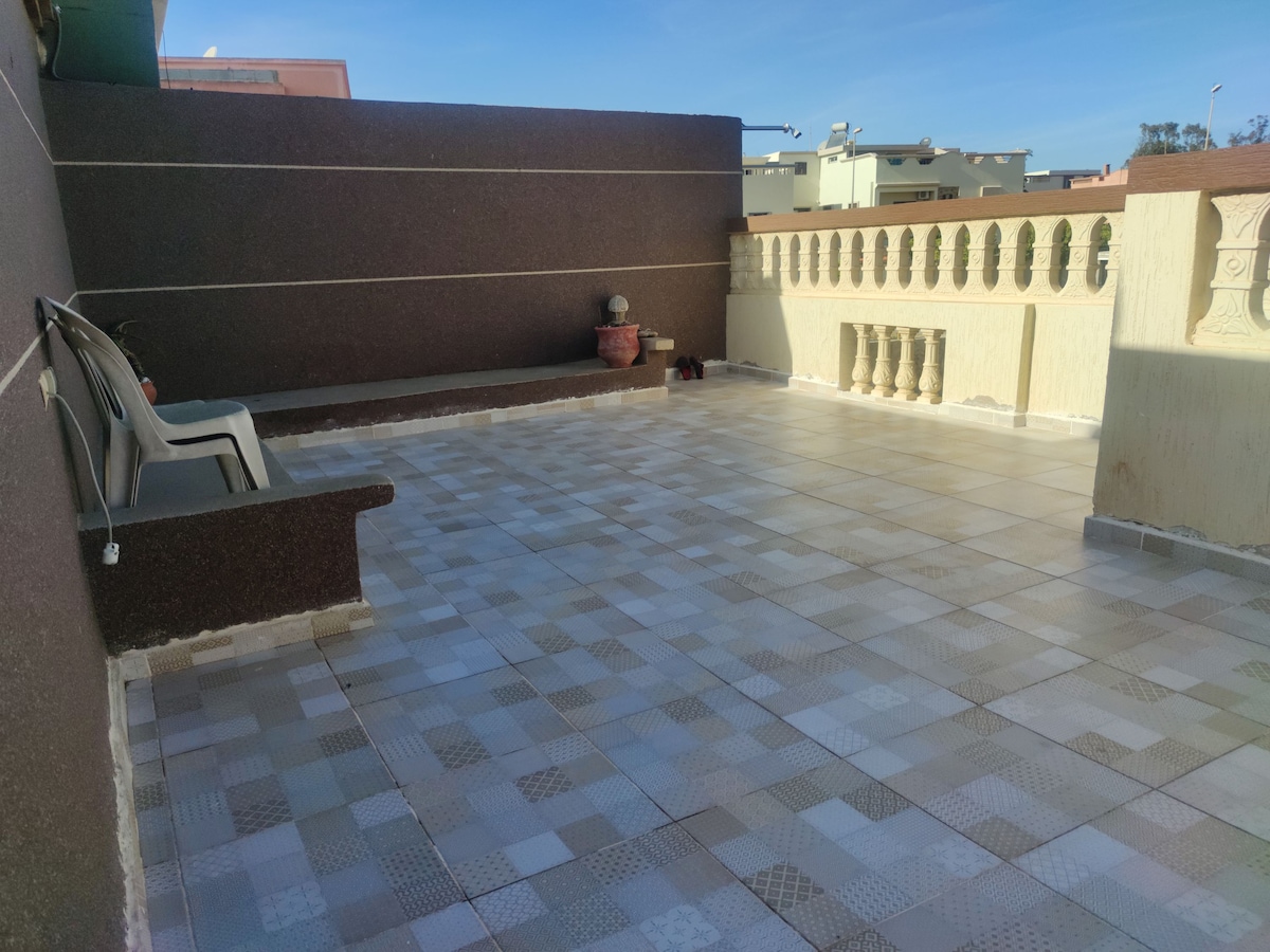 A spacious outdoor terrace is presented, featuring a tiled floor with a variety of muted colors. A few potted plants are positioned around the perimeter, and a pair of simple plastic chairs are visible against the wall, providing a spot for relaxation.
