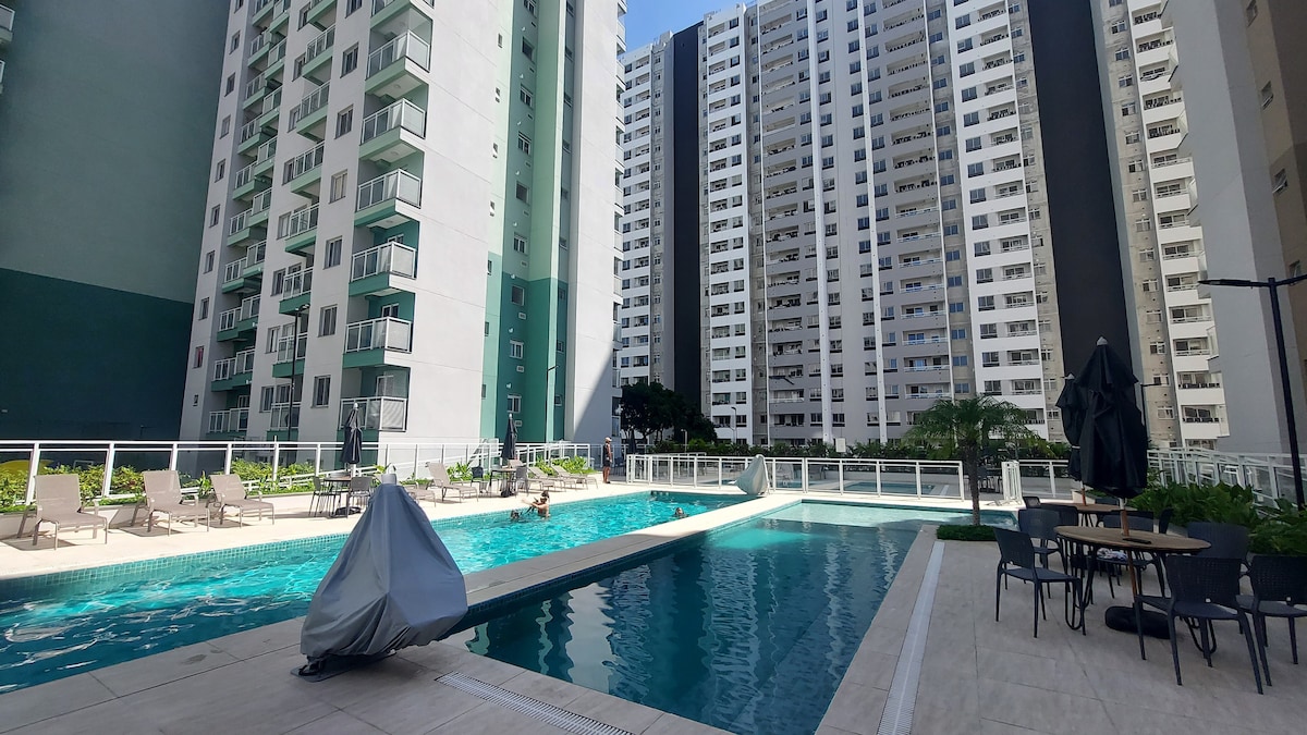 A modern pool area is surrounded by tall residential buildings. This outdoor space features two swimming pools, one with a lane for swimming. Sun loungers are placed along the poolside, and a dining area with tables and chairs is visible nearby.