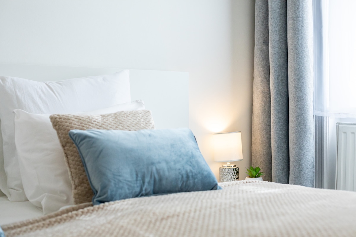A corner of the bedroom showcases a comfortable bed adorned with a mix of textured pillows in neutral and soft blue tones. A softly glowing bedside lamp illuminates the space, while a small potted plant adds a touch of greenery. Light filters in through sheer curtains.