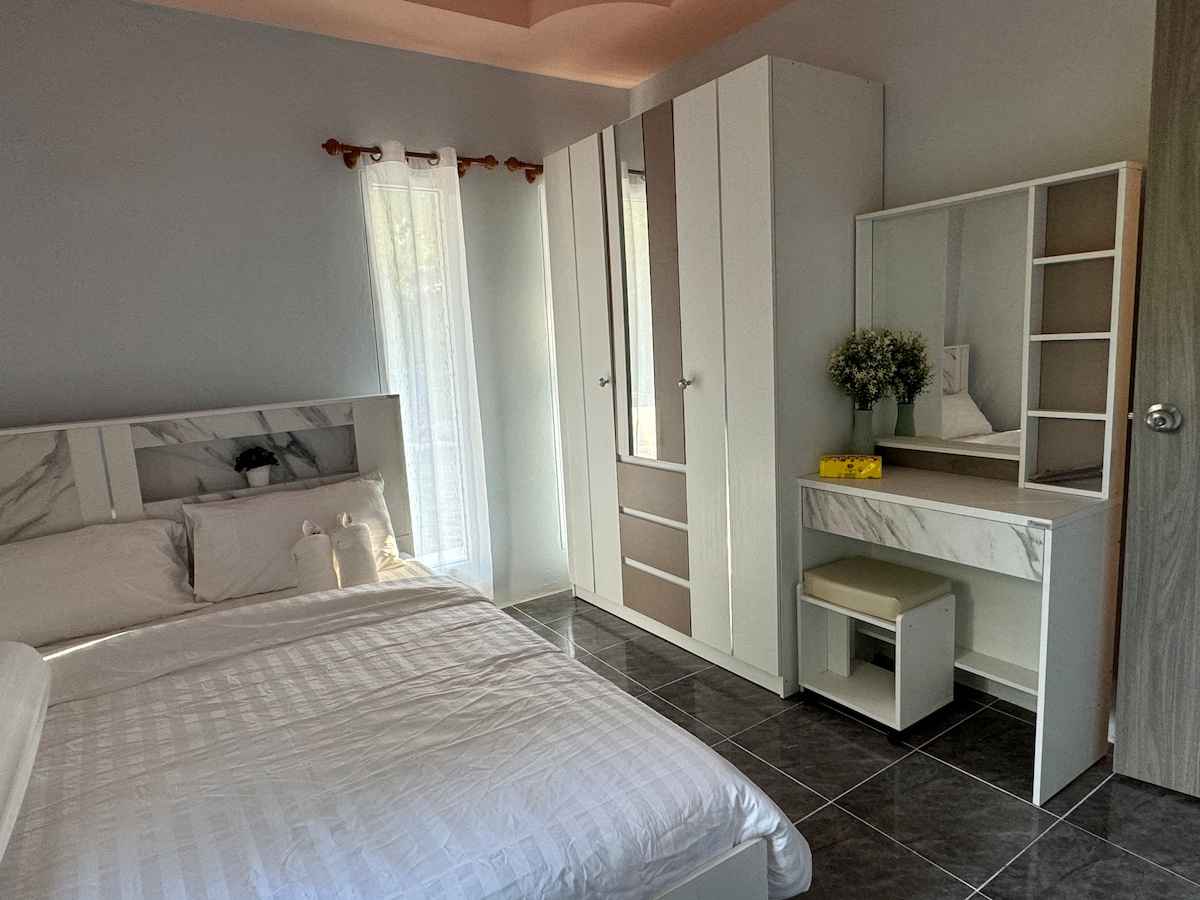 A well-arranged bedroom features a spacious bed with a light-colored coverlet. A modern wardrobe and a neat vanity with a mirror are positioned against the wall. Natural light filters through sheer curtains, enhancing the calming ambiance of the space.