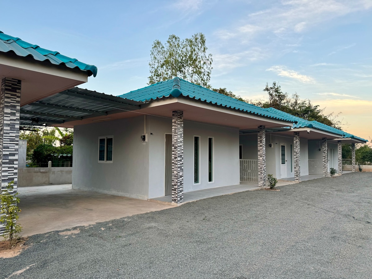 Three identical houses are set in a quiet area, featuring light-colored exteriors and distinctive stone column accents. Each house has a roof in a vibrant turquoise shade. A spacious parking area is visible in front, surrounded by minimal landscaping and a serene atmosphere.