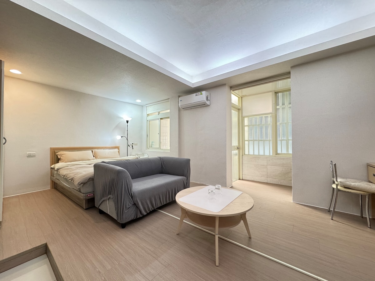 A spacious living area is complemented by a comfortable sofa and a round coffee table. Natural light fills the room through large windows. The inviting sleeping area features a neatly made bed, and light-colored walls enhance the bright ambiance.
