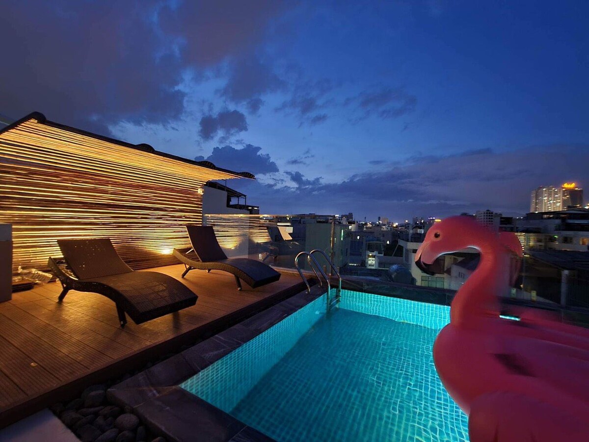 An outdoor rooftop area features a serene pool, illuminated by soft lighting against the evening sky. Lounge chairs are positioned around the pool, with a large flamingo float nearby, creating a relaxed atmosphere for enjoying the city views.