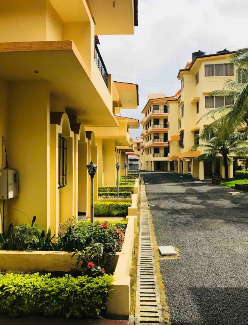 A well-maintained pathway is lined with lush greenery and flowering plants, leading towards a series of yellow, multi-story buildings. Street lamps are positioned along the pathway, enhancing the inviting appearance of the development. The setting reflects a peaceful residential atmosphere.