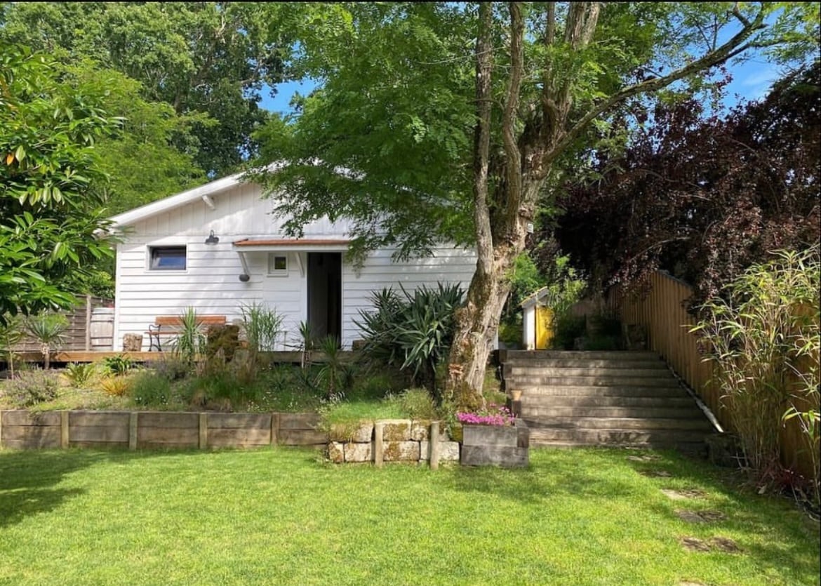 A single-story wooden house is set amidst lush greenery, showcasing a well-maintained garden with various plants. A set of stone steps leads to the entrance, while a bench provides a peaceful sitting area. The façade features simple, clean lines characteristic of modern design.