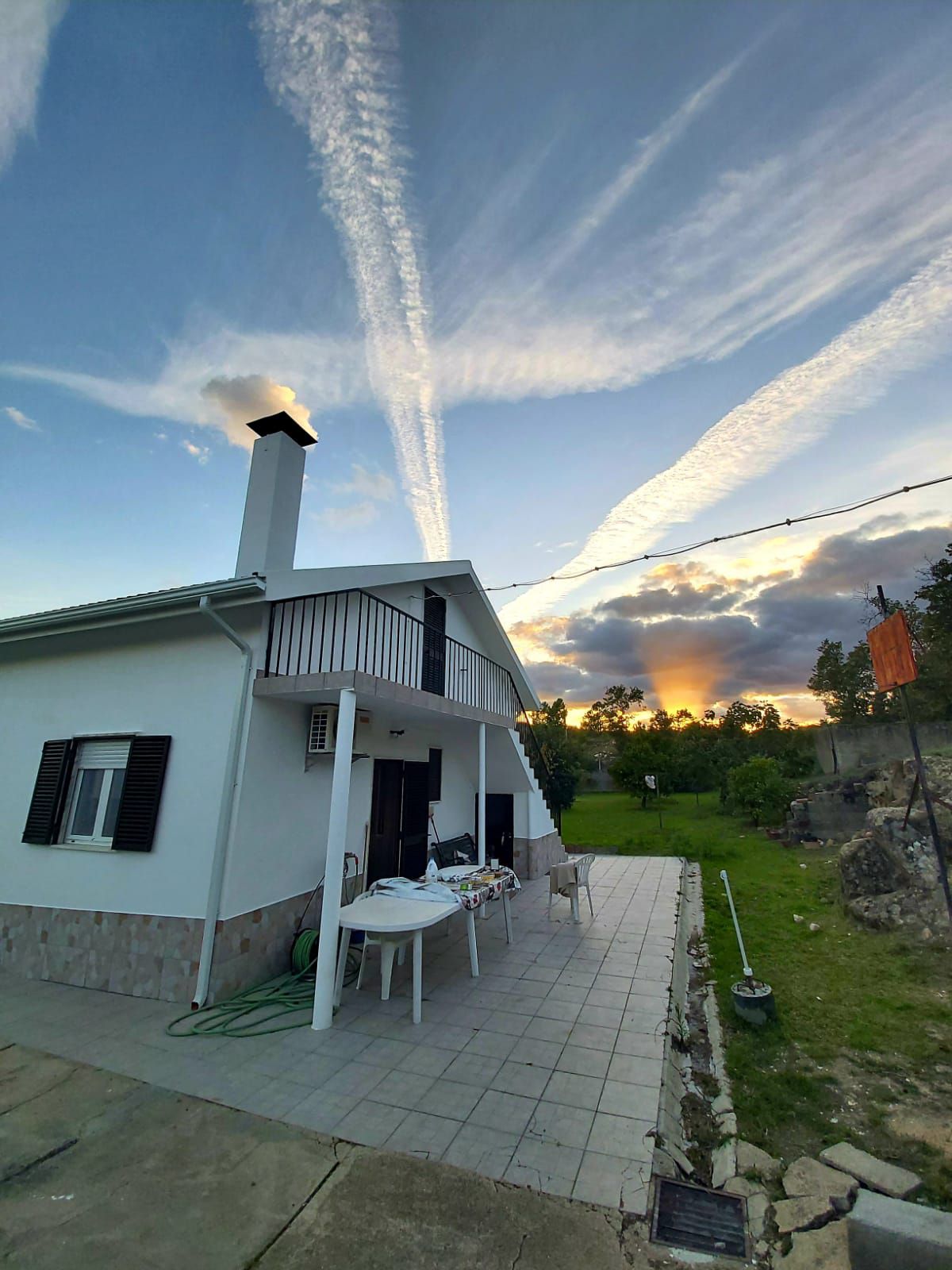 A two-story house is depicted, showcasing a covered patio with a table and chairs. Expansive skies with streaks of clouds create a vibrant sunset backdrop. Lush greenery surrounds the property, enhancing the outdoor space. A chimney rises from the side of the house.