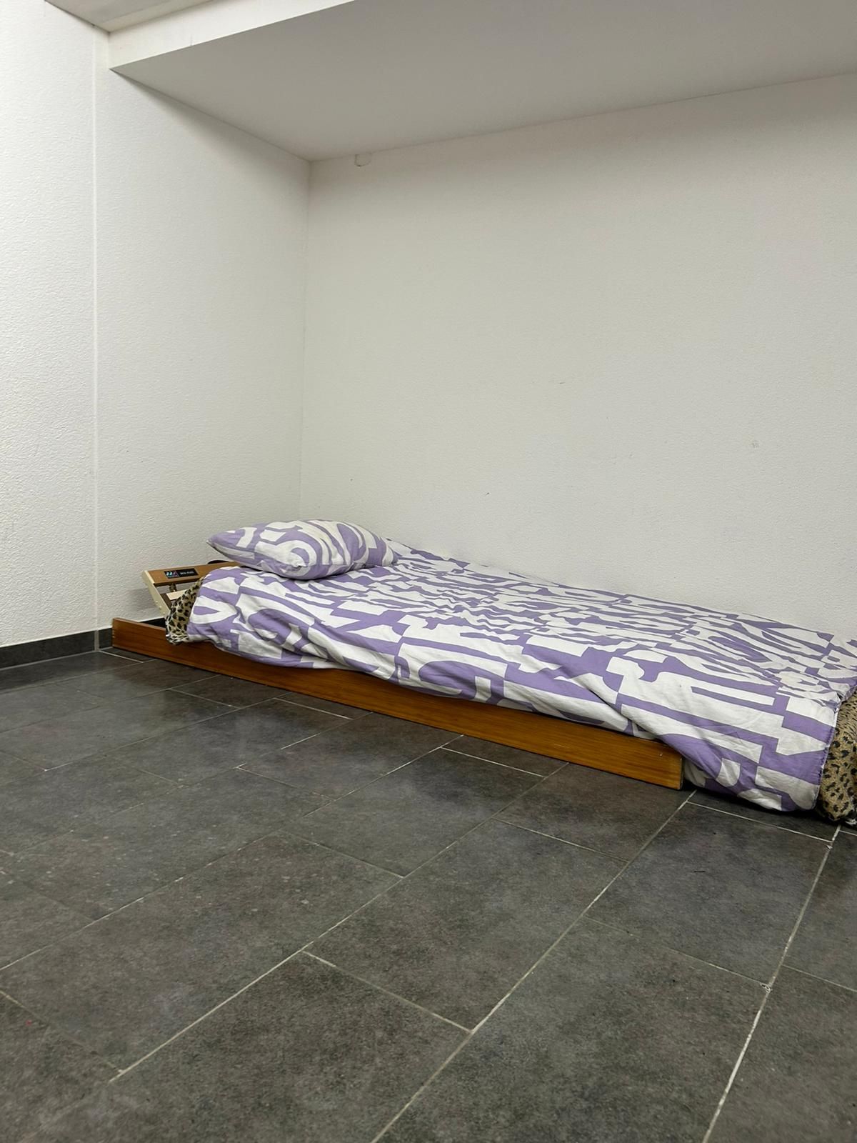 A single bed is positioned against a white wall, featuring a patterned comforter in shades of purple and white. A small shelf is placed beside the bed, providing additional storage. Dark tiles cover the floor, enhancing the room's minimalistic design.