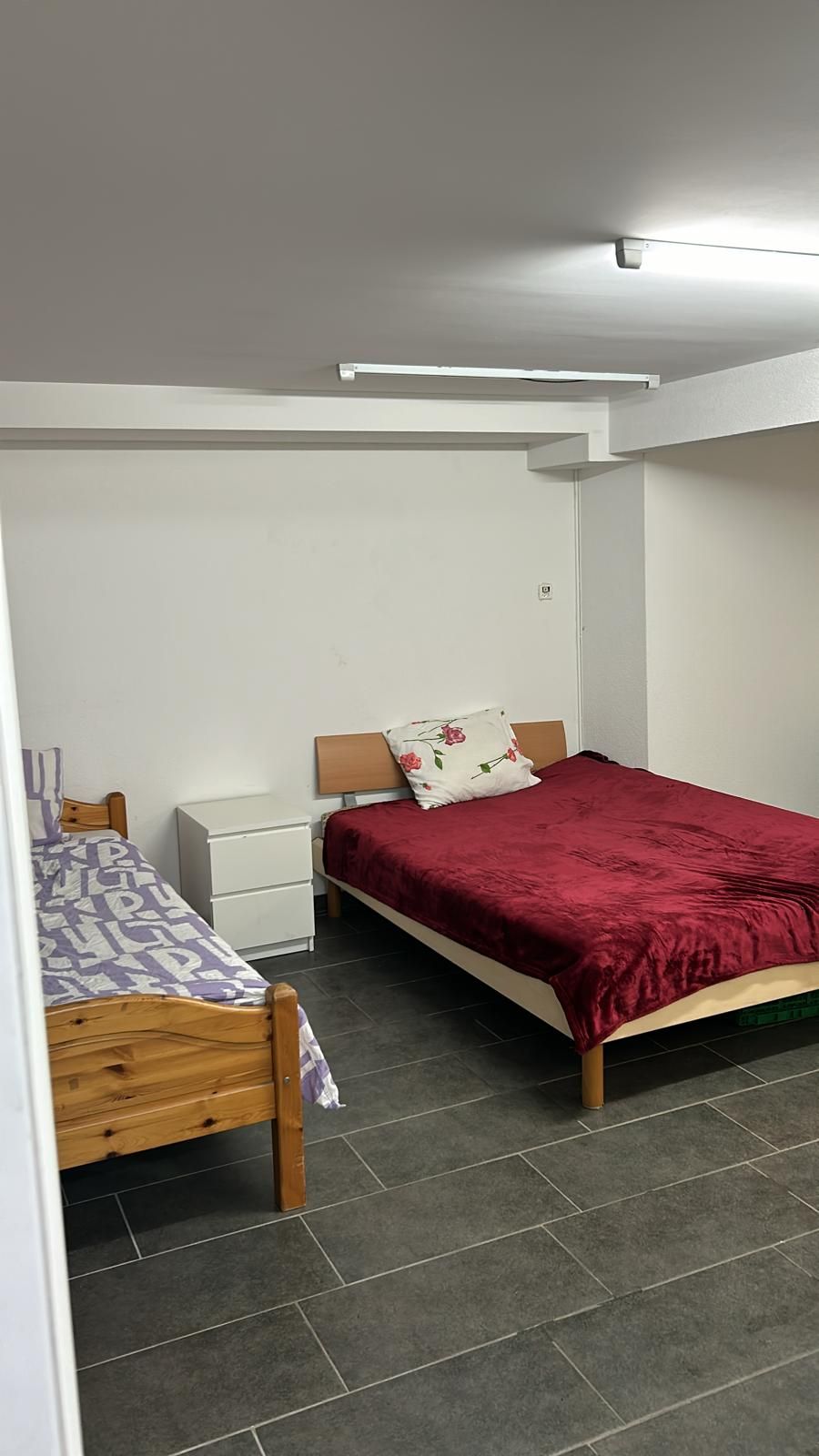 A spacious room is equipped with two distinct beds, one adorned with a red coverlet and the other featuring a patterned blanket. A small bedside table stands beside the red bed, while the room is illuminated by overhead lighting and finished with dark tiled flooring.