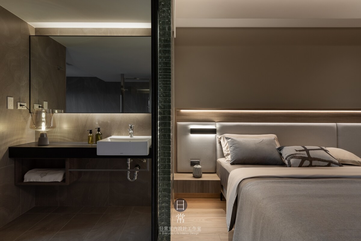 A modern room features a spacious bed with soft bedding and a minimalist design. A sleek bathroom area includes a wall-mounted mirror and a sink. Warm lighting highlights the materials, creating a calm atmosphere throughout the space.
