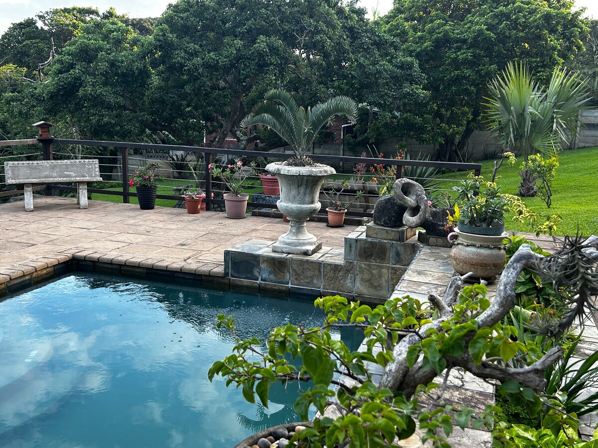 A tranquil outdoor area features a clear pool bordered by a stone patio. Ornamental planters with various plants and flowers are placed around the space. A decorative fountain and a garden view are visible in the background, contributing to a serene atmosphere.