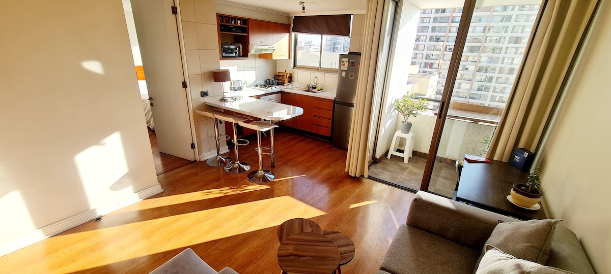 An open living area features a modern kitchenette with wooden cabinetry and a dining bar with three stools. Natural light streams through large windows, illuminating a small desk and the inviting seating area. A balcony is visible, showcasing urban views and a potted plant.