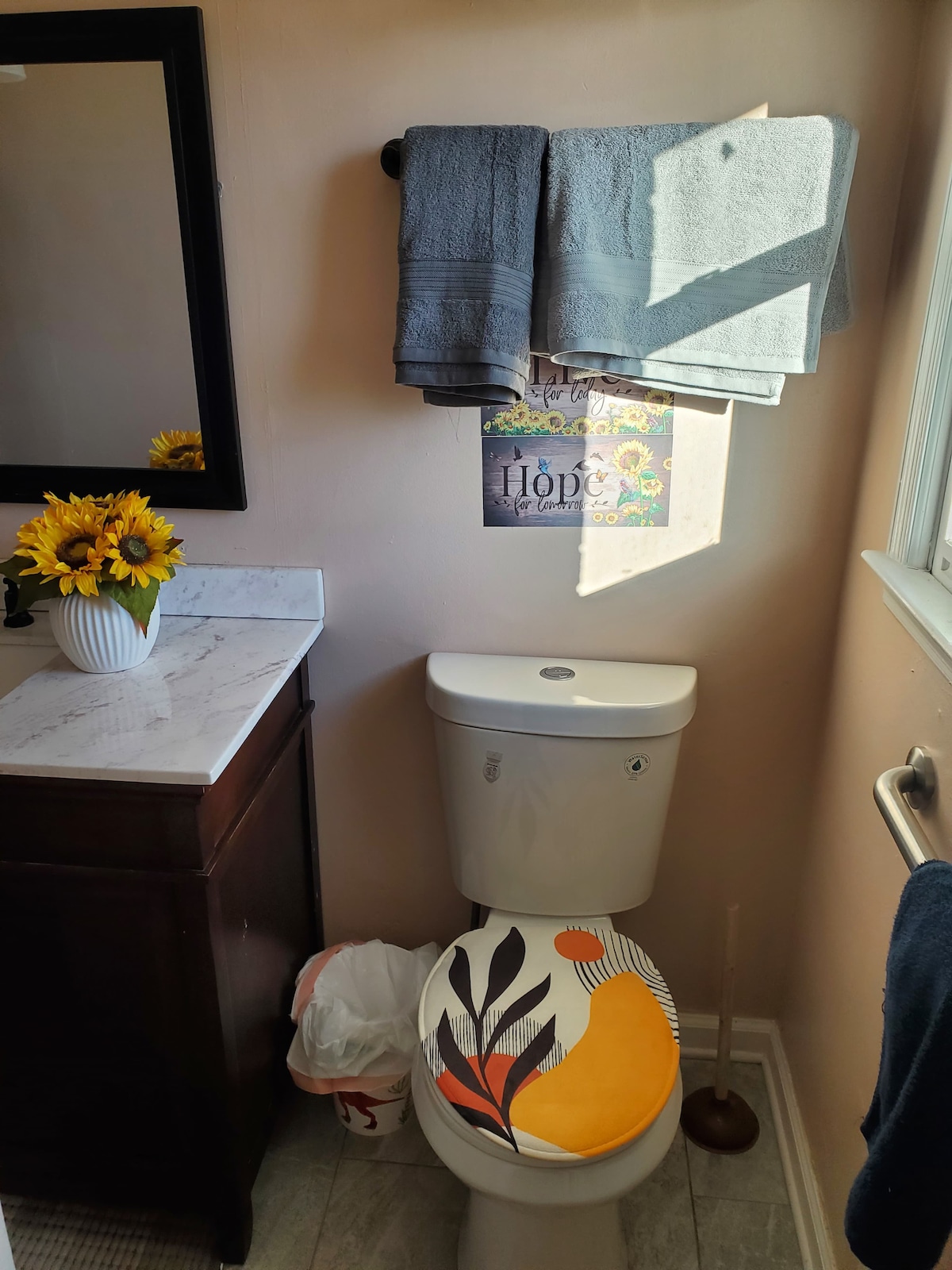 A compact bathroom features a modern white toilet and a dark wood cabinet with a marble countertop. Sunflowers sit in a small vase on the counter. Gray towels are neatly hung above, while a colorful toilet seat cover adds a unique touch to the space.