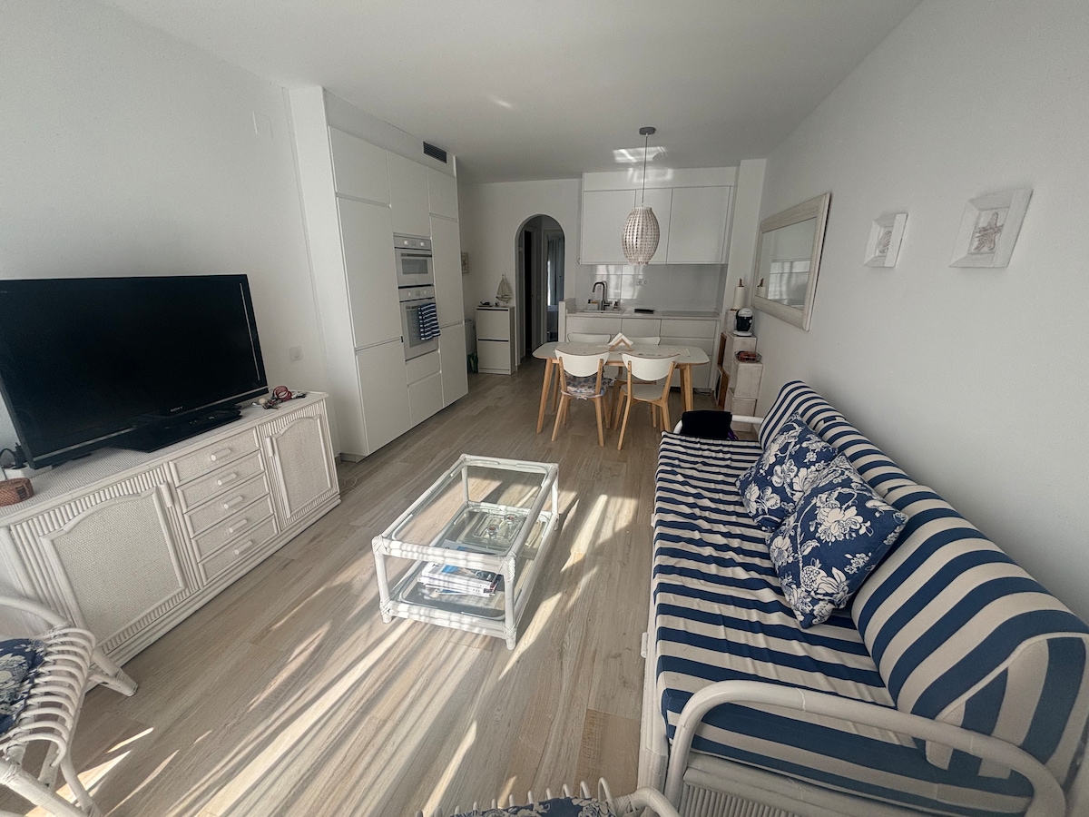 The living area features a comfortable striped sofa paired with a glass coffee table, set against light wooden flooring. A wall-mounted television is positioned opposite, while a modern kitchen is visible in the background, complemented by dining chairs and a pendant light overhead.
