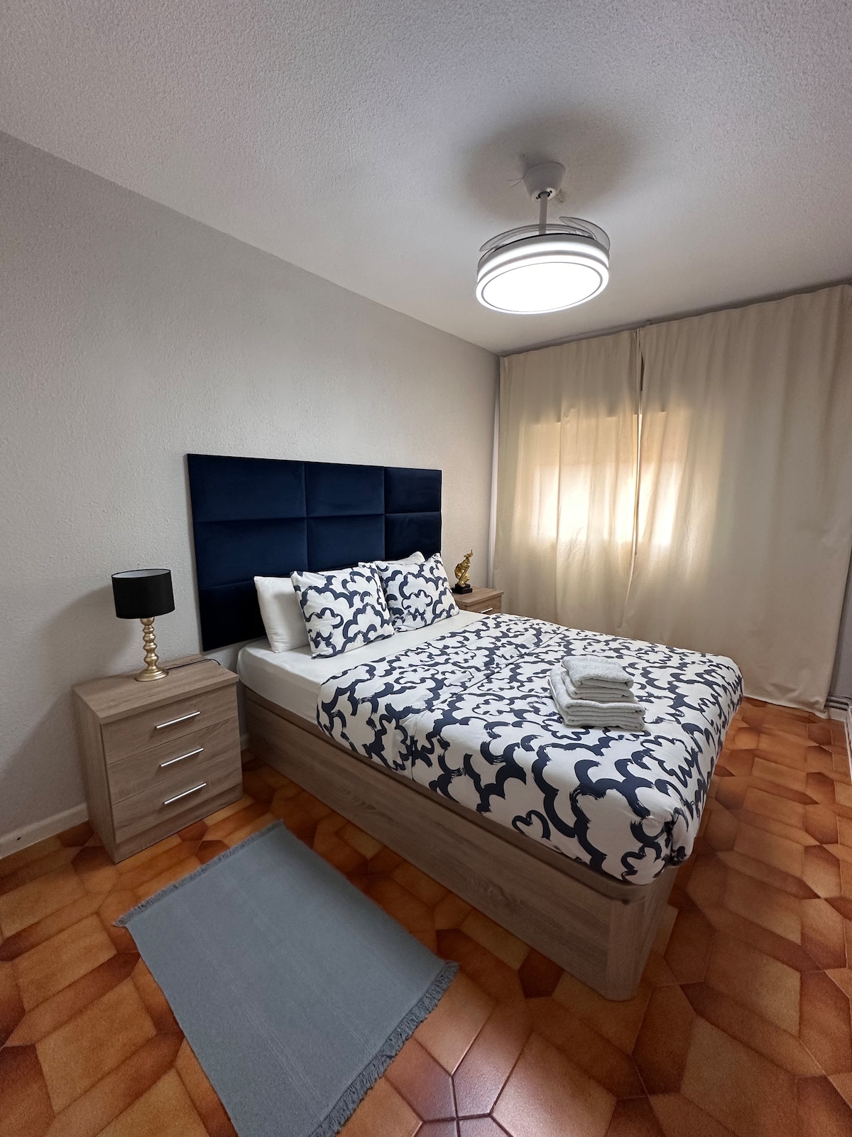 A comfortable bedroom features a spacious bed adorned with a patterned duvet and matching pillows. A bedside table with a lamp stands next to the bed. Light filters through sheer curtains, illuminating the room's warm tile flooring.