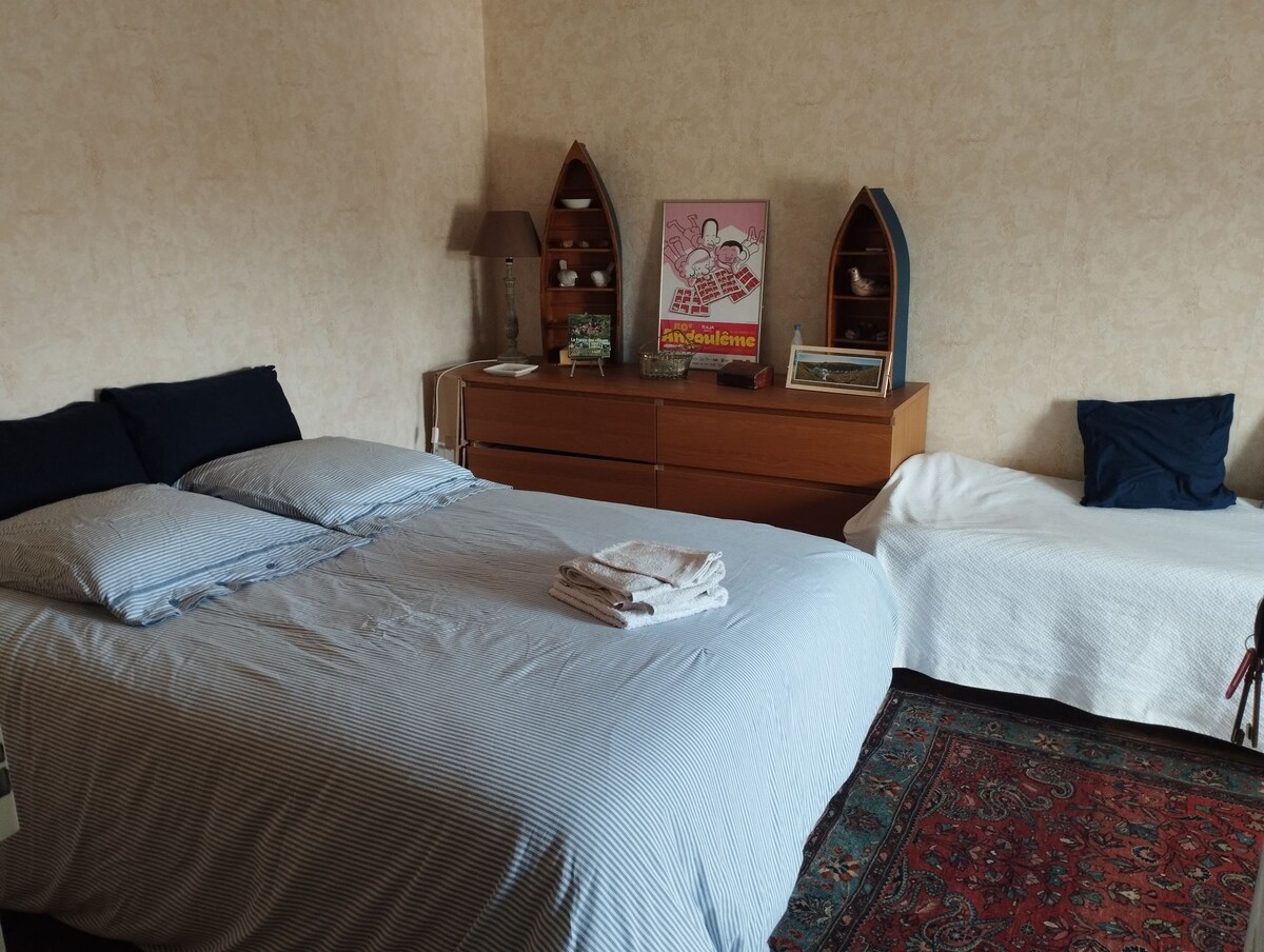 A bright bedroom features a spacious bed with striped bedding and neatly arranged towels on top. A cozy single bed is positioned beside a wooden dresser, adorned with decorative items. Natural light illuminates the room, highlighting the soft tones of the walls and carpet.