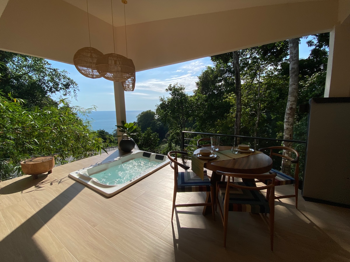 A serene outdoor area is featured, showcasing a private spa and a dining table set for two. The expansive windows overlook lush greenery and the distant ocean, providing a sense of tranquility and connection with nature.