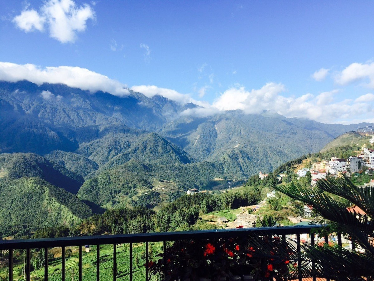 A scenic view of rolling mountains is captured from a balcony. The expansive landscape features a blend of green hills and a blue sky adorned with scattered clouds, highlighting the natural beauty of the surroundings.