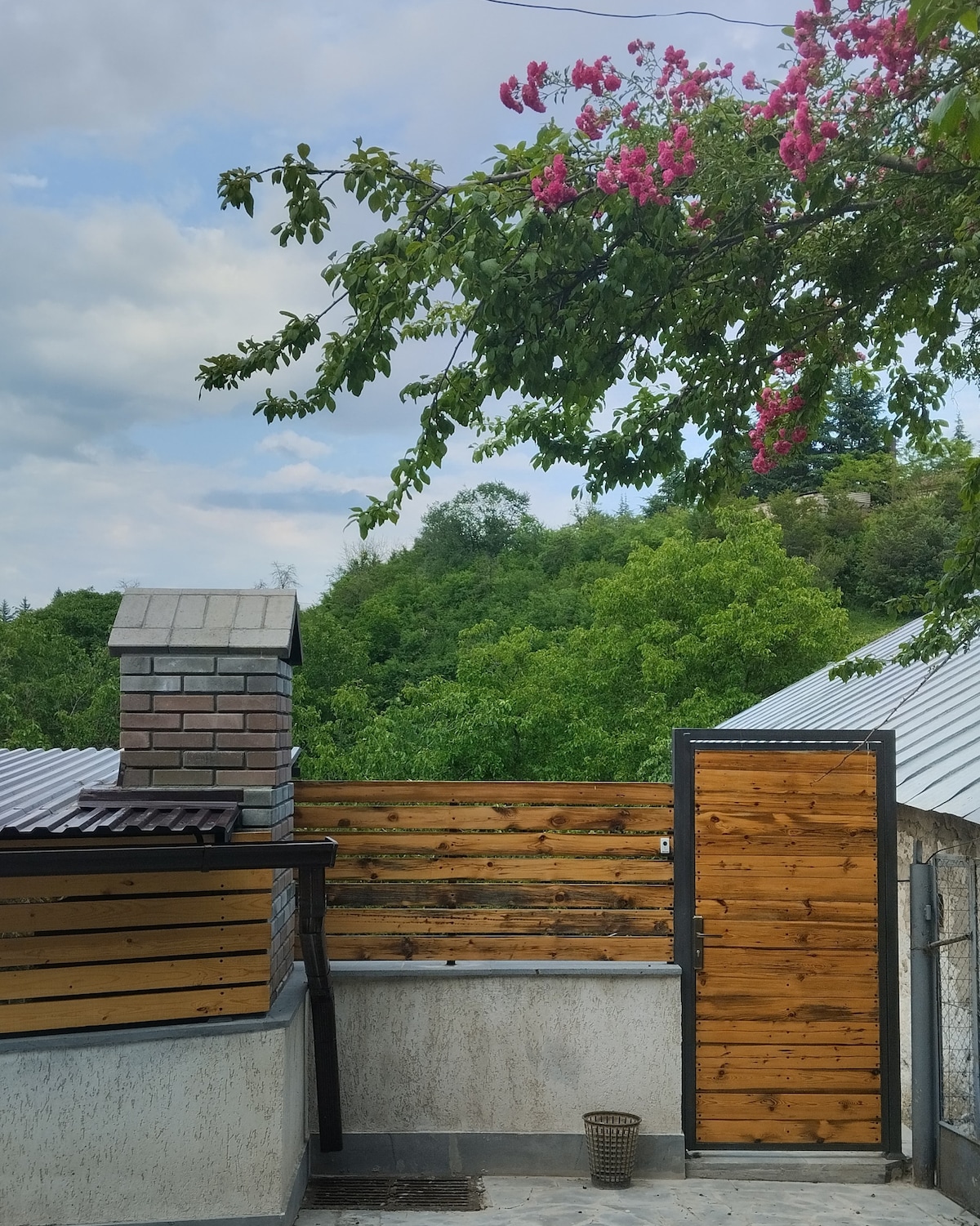 The outdoor area features a wooden fence providing privacy, with a stone chimney visible on one side. Lush greenery can be seen in the background, complemented by flowering branches that add a touch of color to the serene setting.