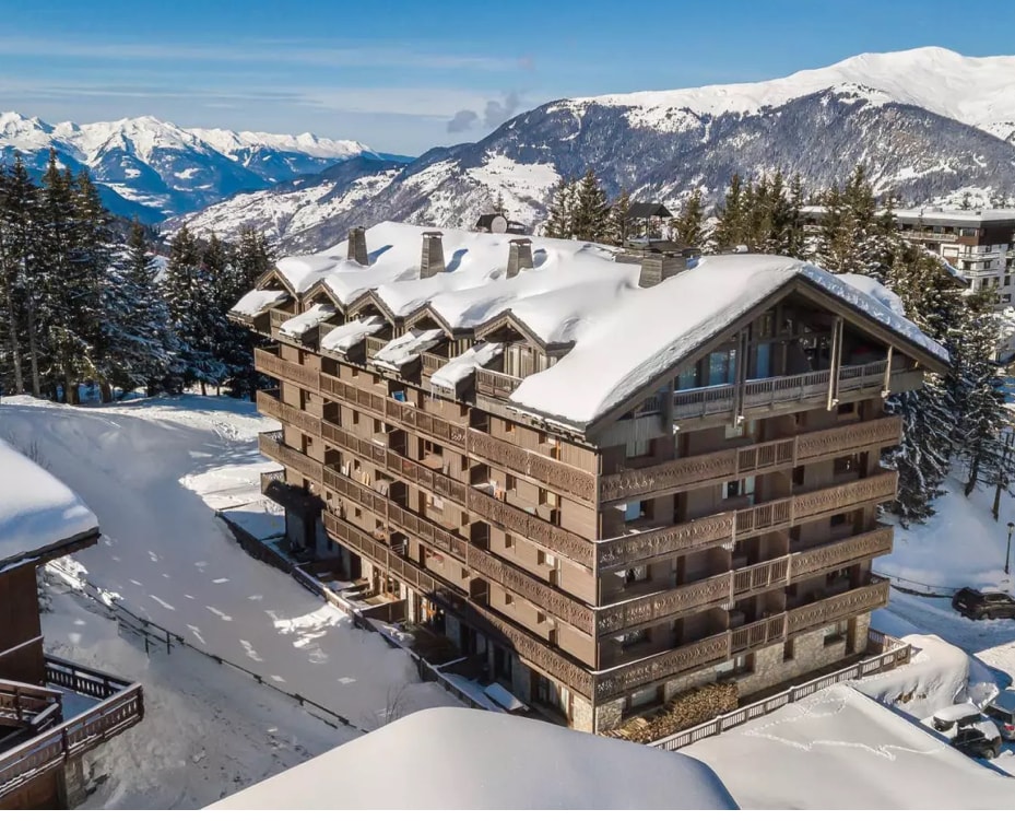 The building's exterior features a mountain lodge design, characterized by wooden beams and stone accents. Snow blankets the rooftop and surrounding pathways, creating a serene winter landscape. The backdrop includes distant snow-capped mountains and clusters of trees, emphasizing the tranquil setting.