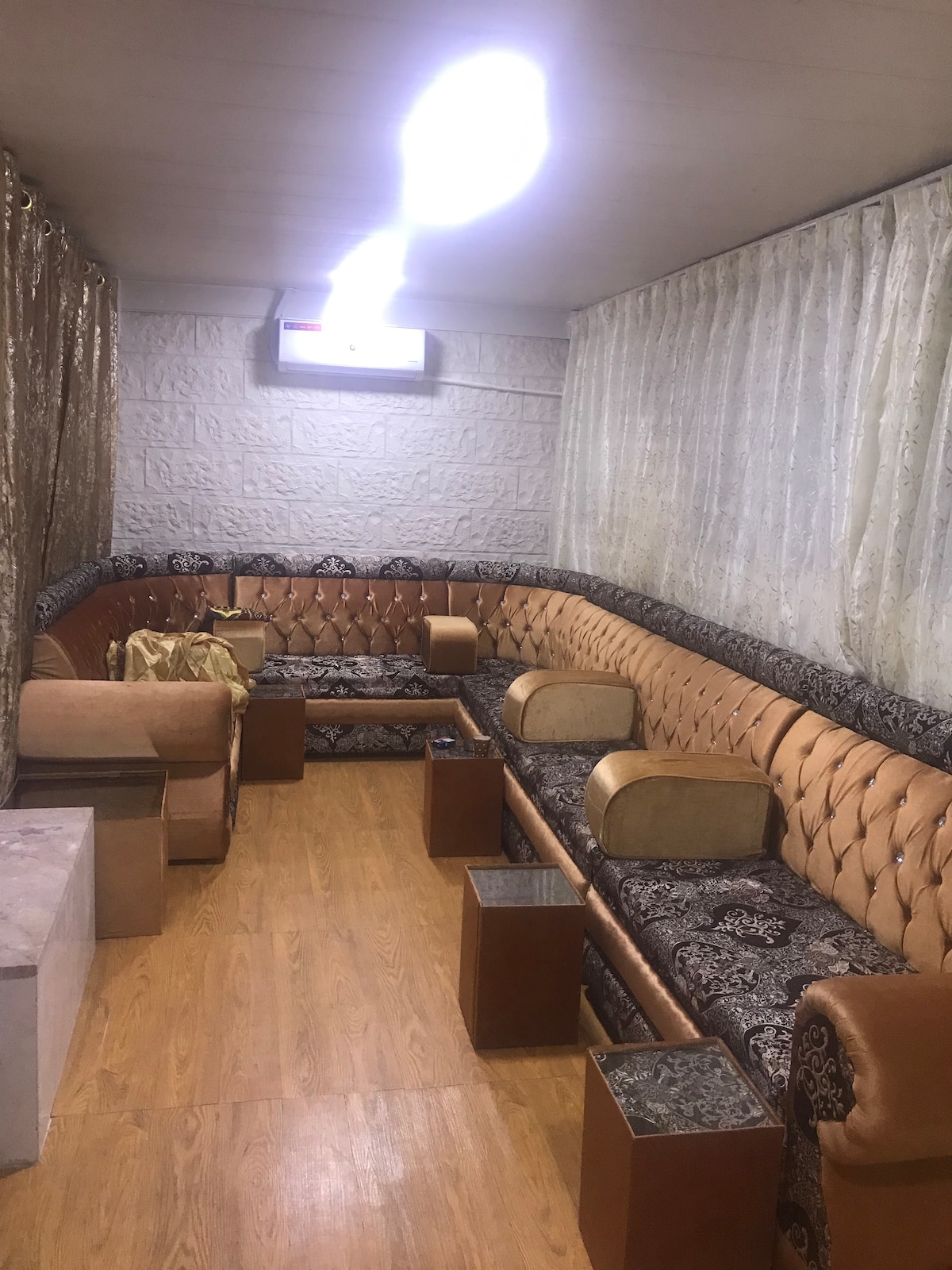 A cozy seating area features a curved brown leather couch adorned with intricate patterns. Small, square wooden tables complement the seating. Soft lighting accentuates the warm wooden floor and light-colored curtains that frame the space, providing a comfortable atmosphere for relaxation.