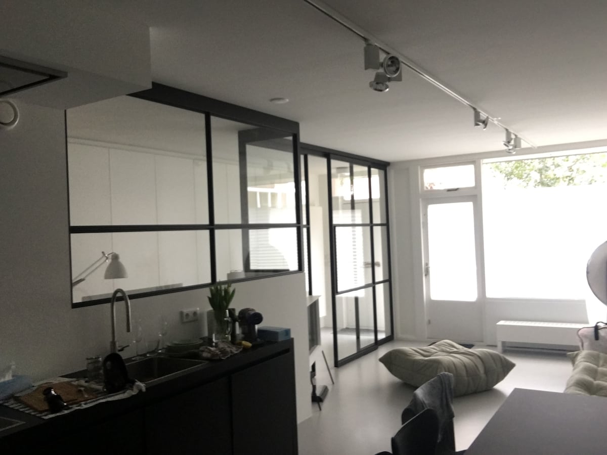 A modern kitchen area is visible, featuring a black island with seating and a sleek sink. Large glass panels allow natural light to flood the space, showcasing a light-filled sitting area with soft furnishings arranged on the floor nearby.