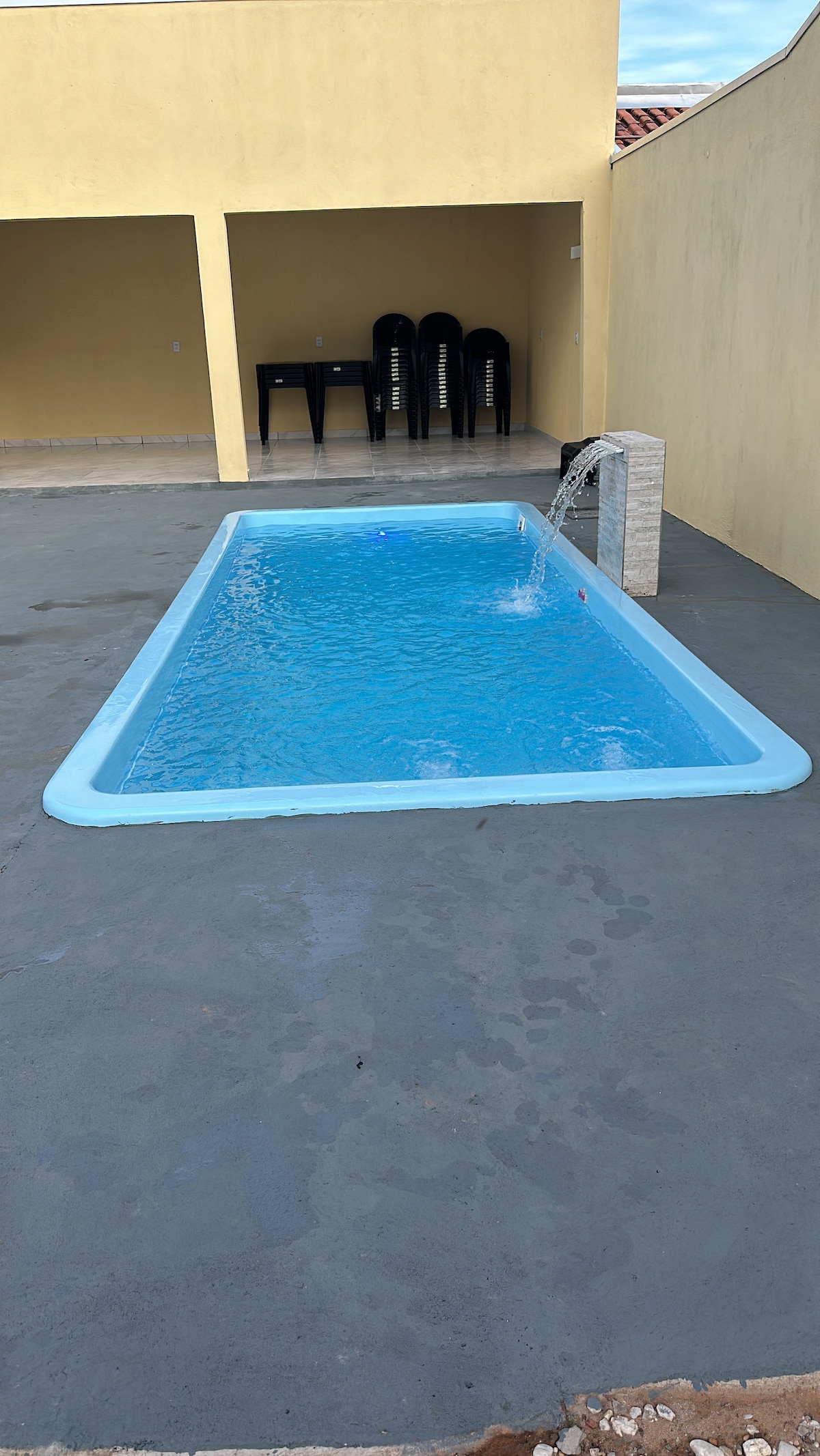 A rectangular swimming pool is featured, surrounded by a concrete patio. A small water feature is visible, gently cascading water into the pool. In the background, a shaded area with several black chairs can be seen, providing a space for relaxation.
