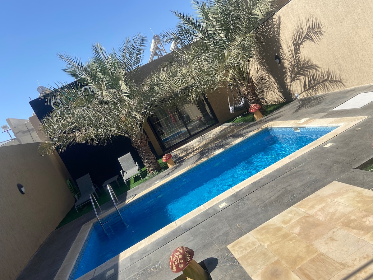 A private pool is surrounded by palm trees and a stone patio. Comfortable seating areas are positioned nearby. Neutral-toned walls provide a serene backdrop. The pool features clear blue water, inviting for a refreshing swim under bright sunlight.