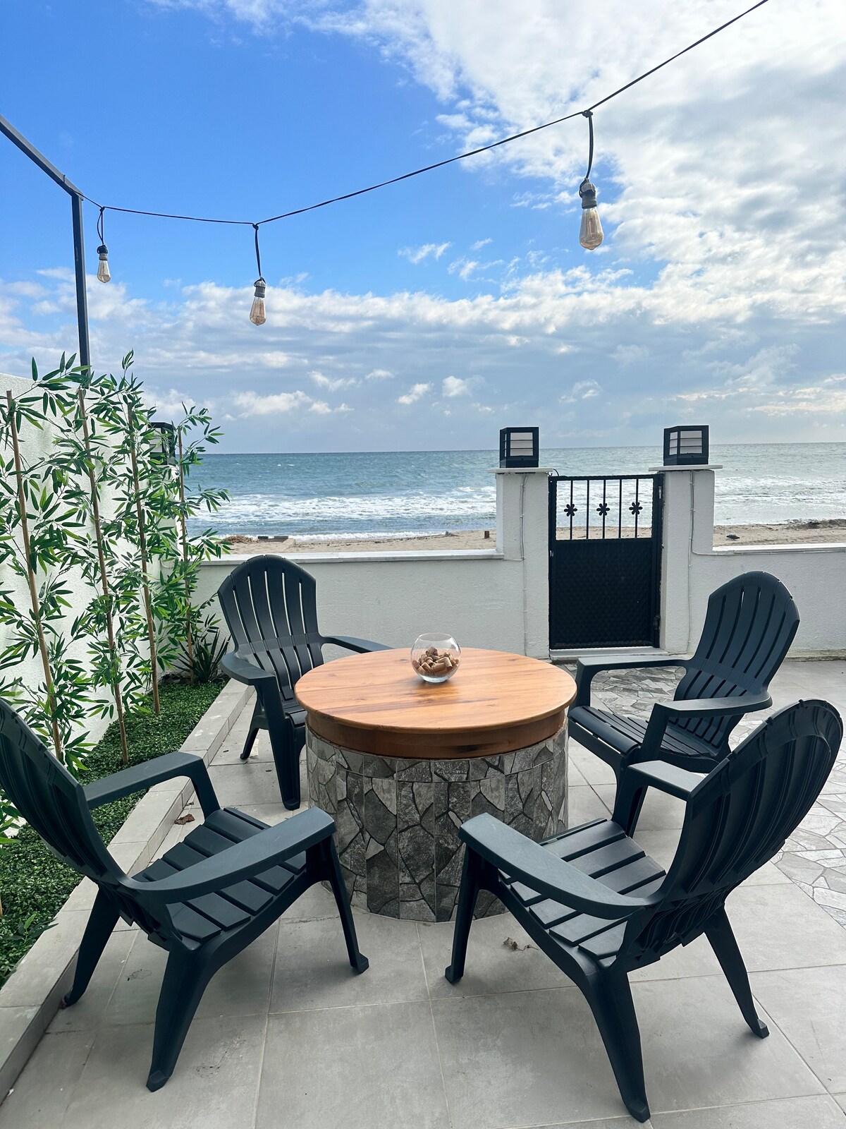 A cozy outdoor seating area features a circular stone table surrounded by four dark green chairs. String lights are hung above, creating a warm ambiance. Waves can be seen in the background, providing a serene view of the nearby beach.