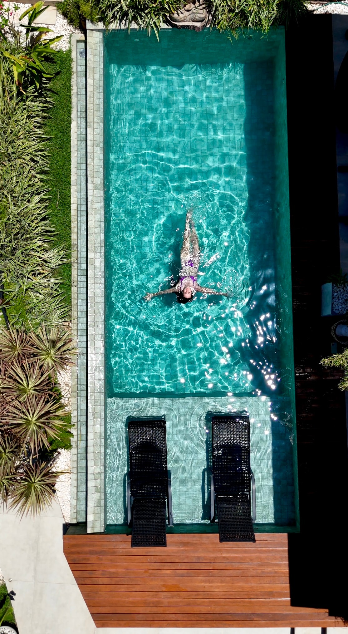 An aerial view captures a clear blue pool bordered by lush greenery. Two black lounge chairs are positioned beside the pool, while a person swims leisurely, creating ripples in the water. The pool area is complemented by wooden decking and a variety of plants.