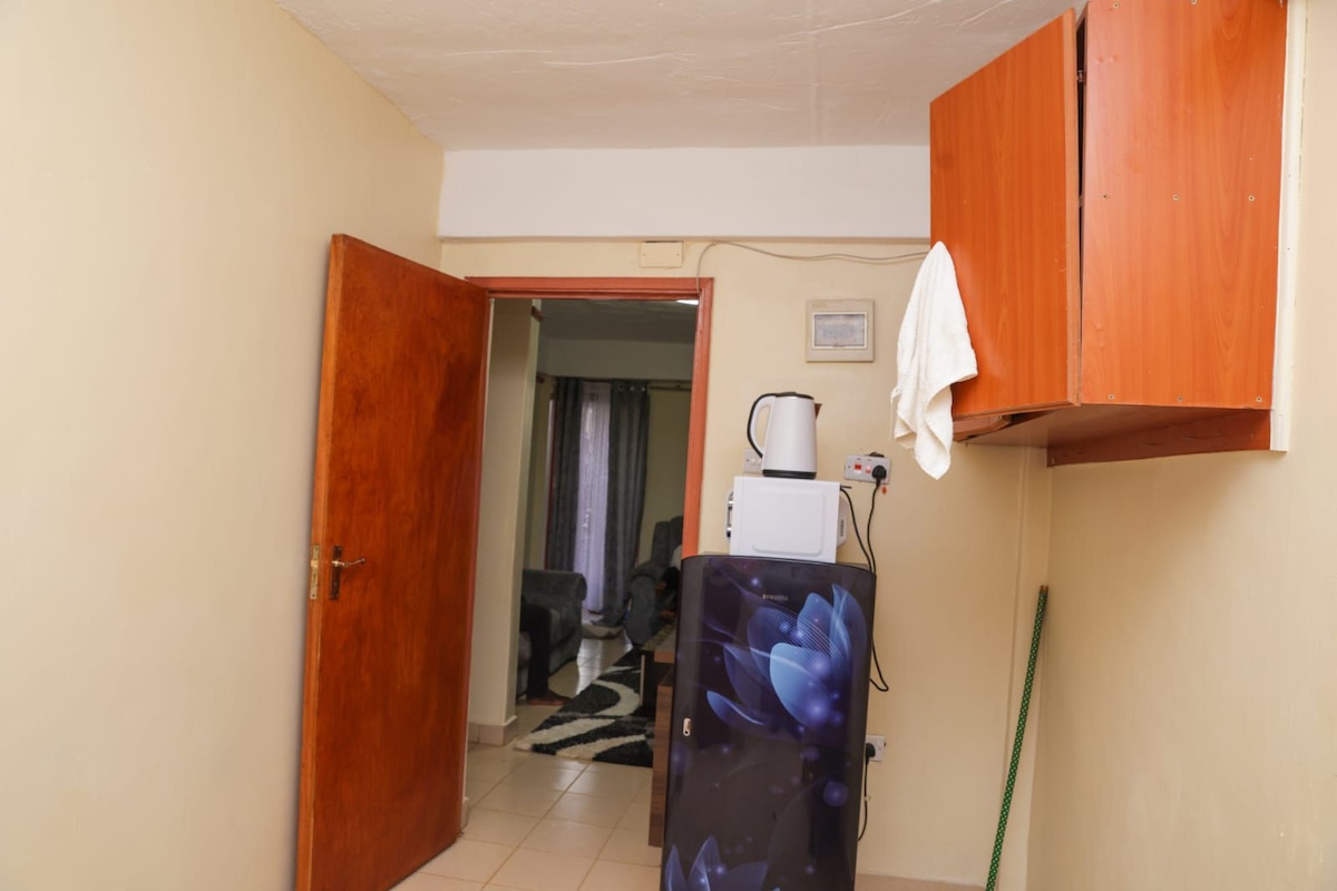 The image depicts a small kitchen area with a wooden cabinet mounted above a countertop. A kettle and microwave are positioned on the countertop, adjacent to a door leading to another room. The floor tiles are light-colored, and a rug is visible in the room beyond.