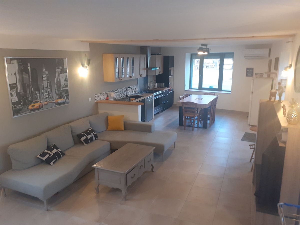 A spacious living area features a comfortable sectional sofa facing a coffee table. An adjacent dining table surrounded by chairs is positioned near a window, allowing for natural light. Modern kitchen appliances and cabinetry are visible, with a large cityscape artwork adorning the wall.