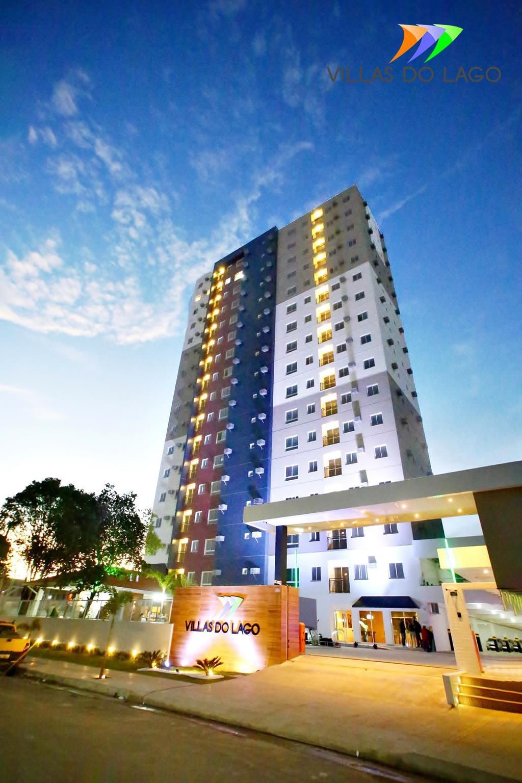 A modern multi-story building is showcased against a backdrop of a twilight sky. Illuminated windows create a glow, highlighting the architectural design. The entrance features a sign, and the landscape includes a parking area and trees, contributing to the overall ambiance.