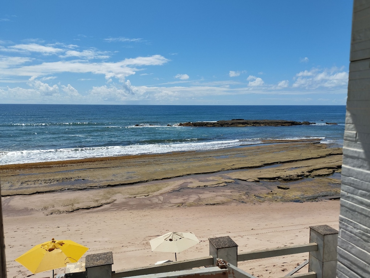 A clear view of the coastline is presented, showcasing gentle waves lapping at the shore. Two yellow umbrellas offer shade on the sandy beach, while rocky formations are visible in the water, creating a natural contrast with the blue sea and sky.