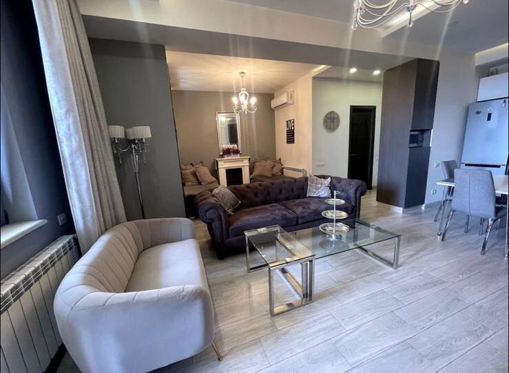A living area is presented with a plush dark sofa and a modern light-colored couch. A glass coffee table sits in the center, surrounded by soft armchairs. The space is warmly illuminated by a chandelier, while a dining area is visible in the background.