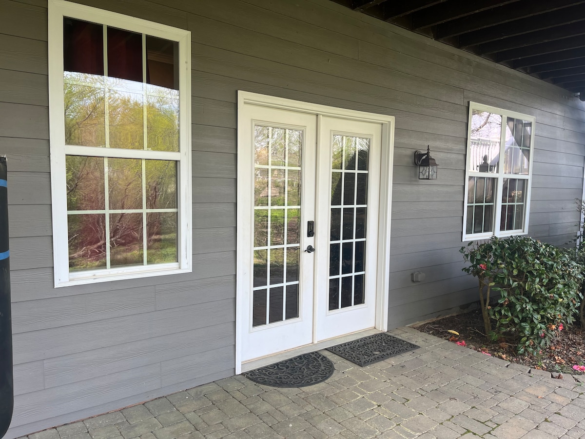 The entrance features a set of double doors with glass panels, framed by two large windows on either side. The stone pathway leads to the door, bordered by low greenery and a well-maintained landscape, inviting guests to the private apartment.