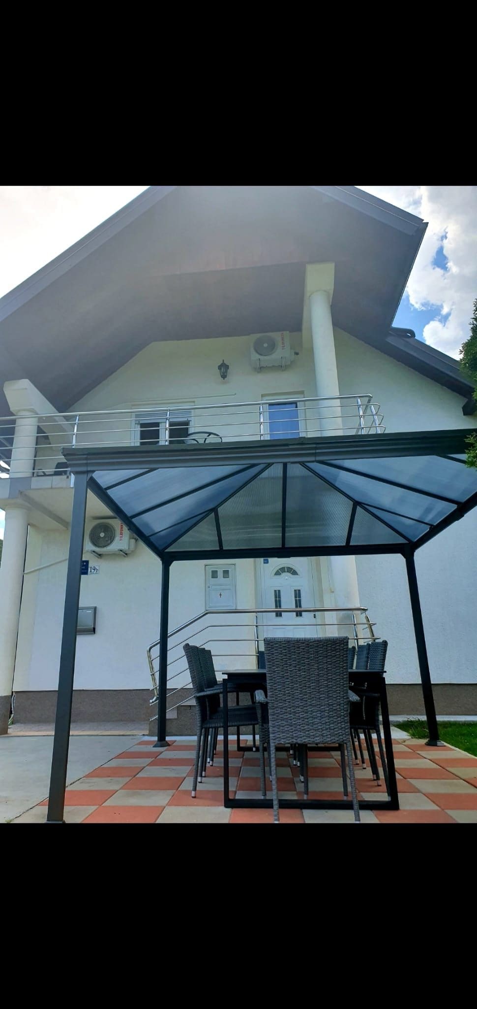 An outdoor dining area features a large gazebo, providing shade over a set of six chairs and a rectangular table. The backdrop showcases a two-story white house with a modern design, complemented by a well-maintained exterior.