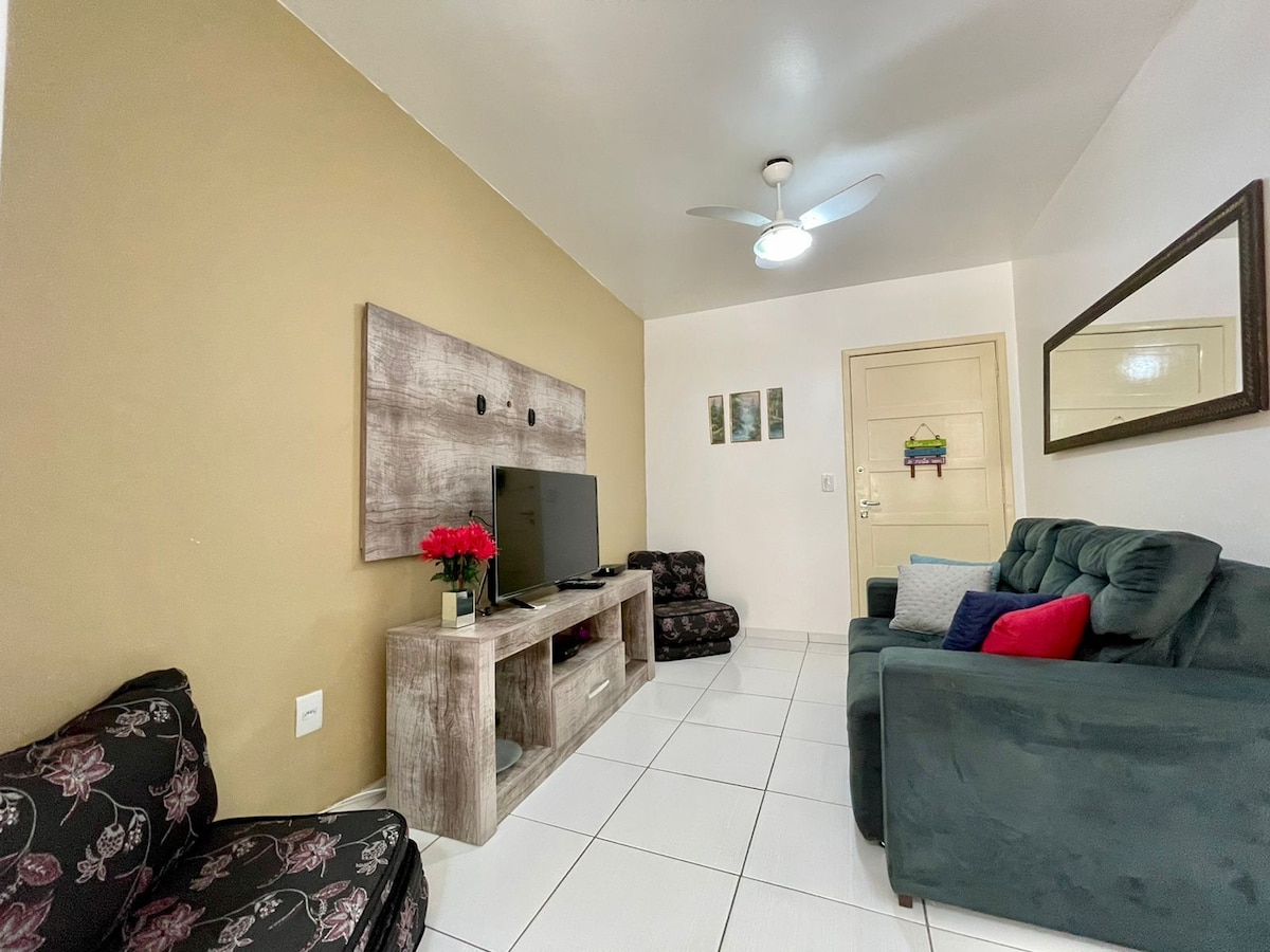 A cozy living area is presented with a dark green couch and patterned armchairs. A TV is mounted on a wooden panel against a light, warm-toned wall. A flower arrangement sits on a low wooden unit, with a ceiling fan above providing added comfort.