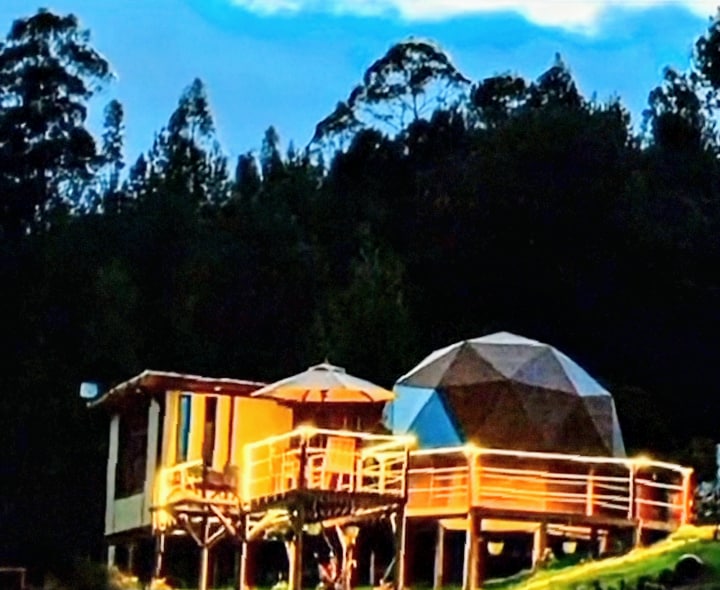A modern geodesic dome is seen next to a cozy wooden structure, both surrounded by lush greenery. A spacious deck with a railing is illuminated, enhancing the inviting atmosphere. The backdrop features a scenic view of nature under a twilight sky.